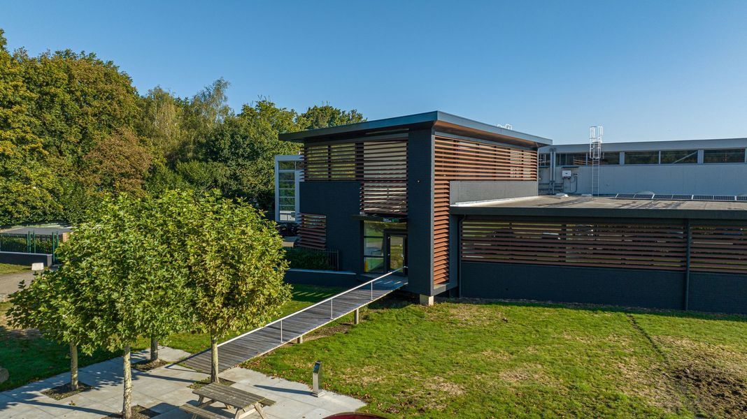 Modern gebouw met houten lamellen, grote ramen en een grasveld naast bomen onder een blauwe hemel.