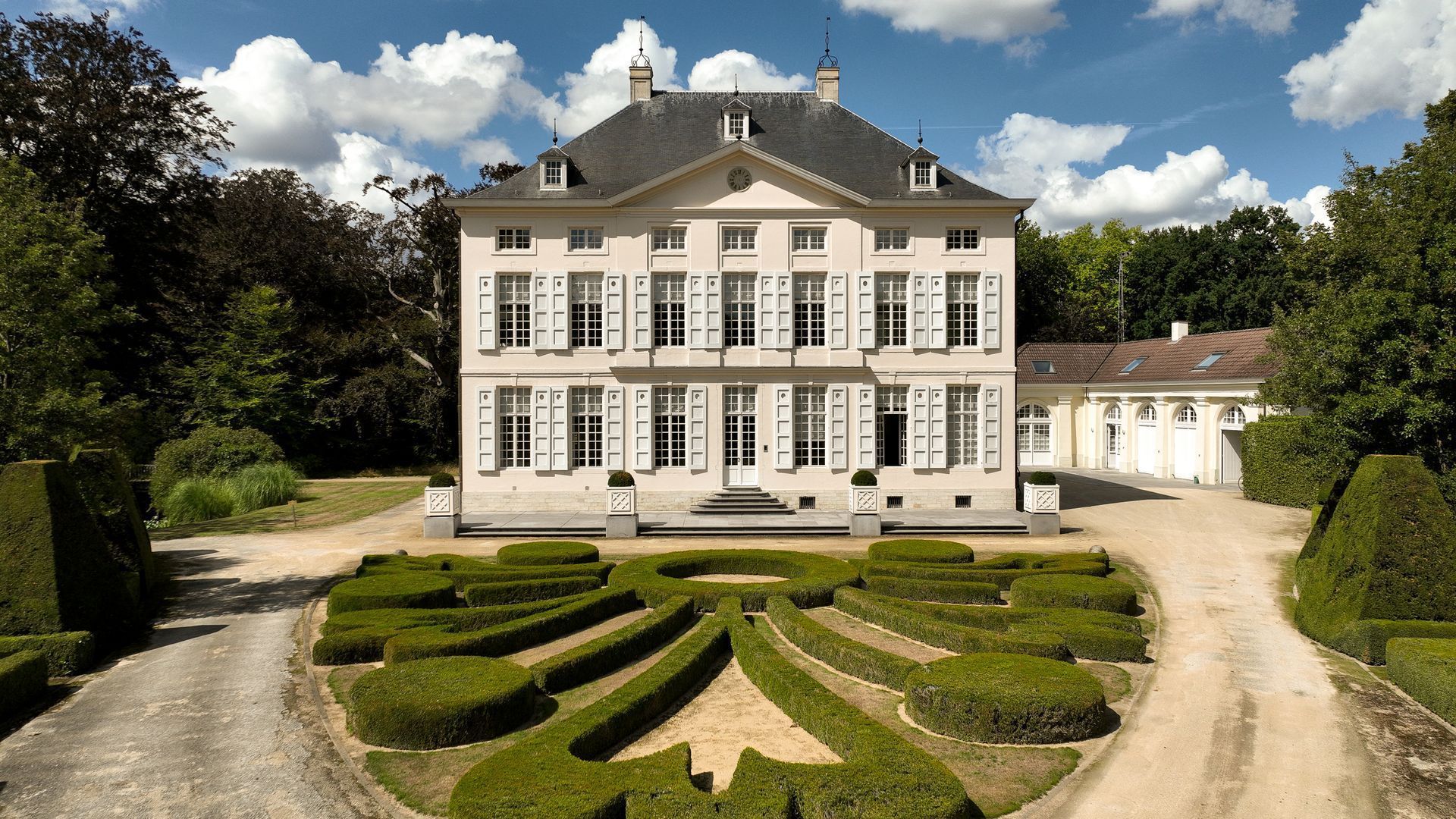 Groot wit landhuis met een keurig onderhouden tuin en een grindpad onder een blauwe hemel.