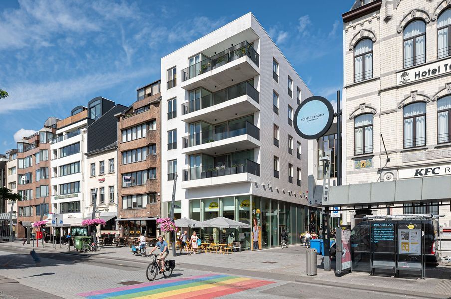 Stadsstraat met moderne appartementen, fietsers en een regenboogzebrapad onder een blauwe hemel.