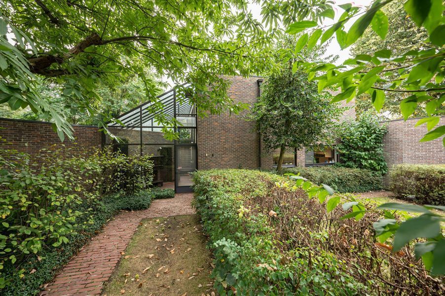 Een tuinpad leidt naar de ingang van een bakstenen huis, onder lommerrijke bomen en hagen.