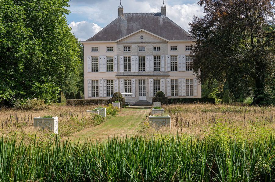 Een groot wit herenhuis met veel ramen, gelegen aan de rand van een grasveld en bomen onder een heldere hemel.