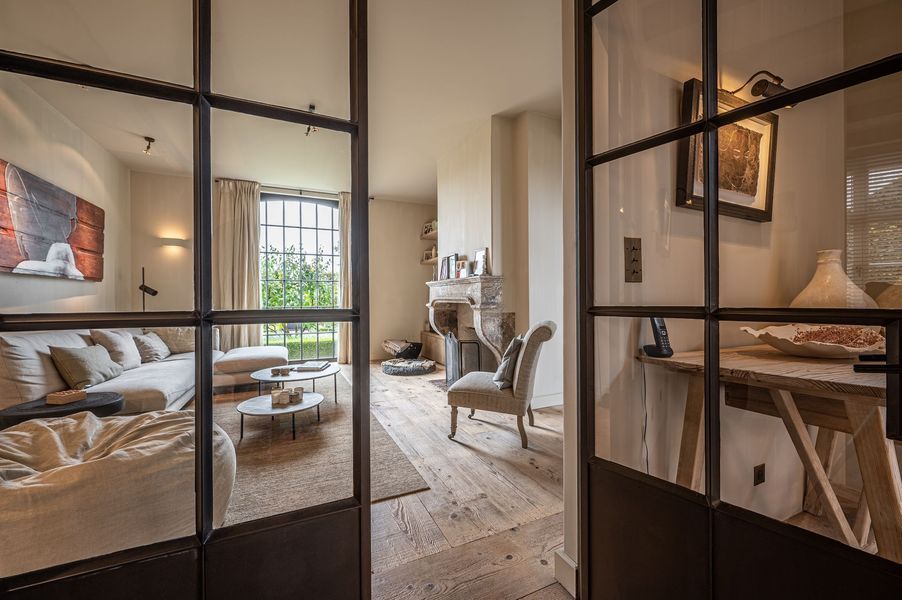 Moderne woonkamer met beige bank, fauteuils, houten vloer en glazen deuren met zwarte kozijnen.