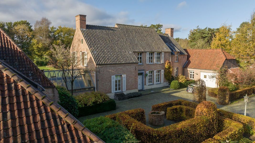 Bakstenen huisje met donker pannendak, tuinhagen en herfstbomen in een zonnige, landelijke omgeving.