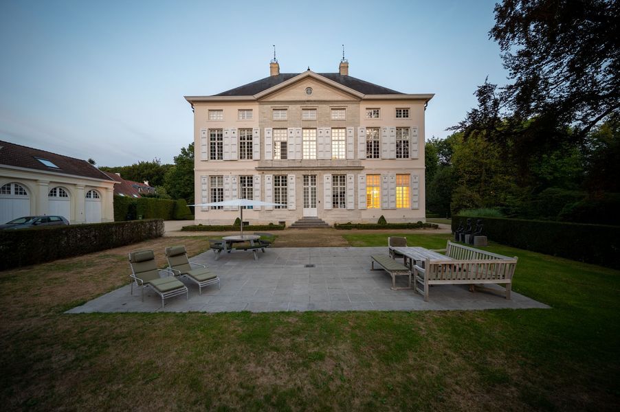 Groot, crèmekleurig herenhuis met verlichte ramen, terrasstoelen en een gazon in de schemering.