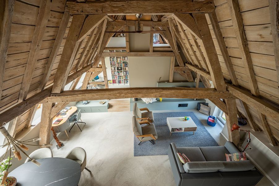 Loftinterieur met zichtbare houten balken, bank, stoelen, tafel en uitzicht op een lager gelegen kamer door de open vloer.