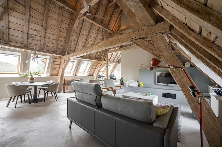 Moderne zolderwoonkamer met zichtbare houten balken, grijze bank, eettafel en grote ramen.