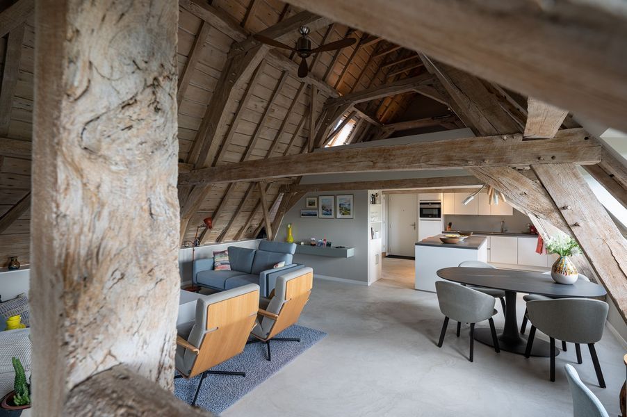 Rustieke loft-woonkamer met zichtbare balken, grijze banken en een eettafel onder een schuin houten plafond.