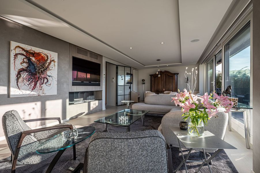 Lichte, moderne woonkamer met grijze banken, glazen salontafel, wanddecoratie en grote ramen met uitzicht op de stad.