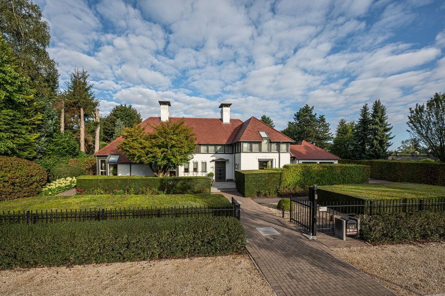 Groot wit huis met rood dak en keurig gesnoeide hagen achter een afgesloten oprit onder een bewolkte hemel.