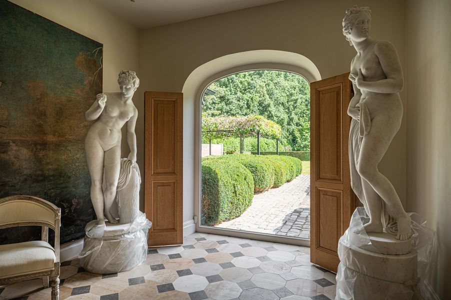Elegante hal met witte beelden, open houten deuren en uitzicht op een verzorgde tuin.