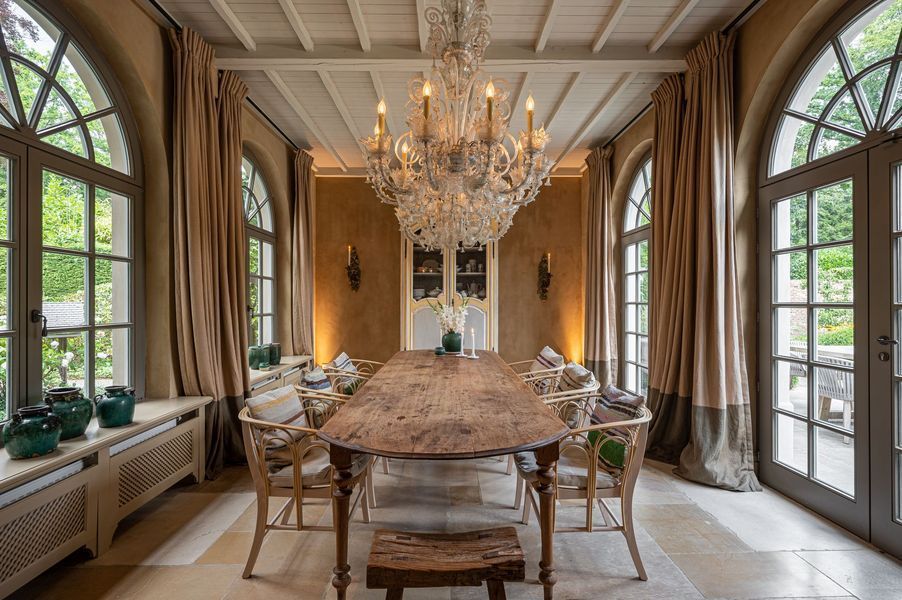 Elegante eetkamer met kroonluchter, lange houten tafel, boogvensters en gedrapeerde gordijnen.