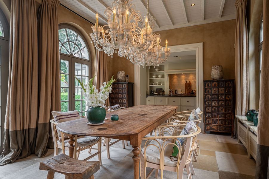 Elegante eetkamer met een kroonluchter, een lange houten tafel, een boogvormig raam en een neutrale inrichting.