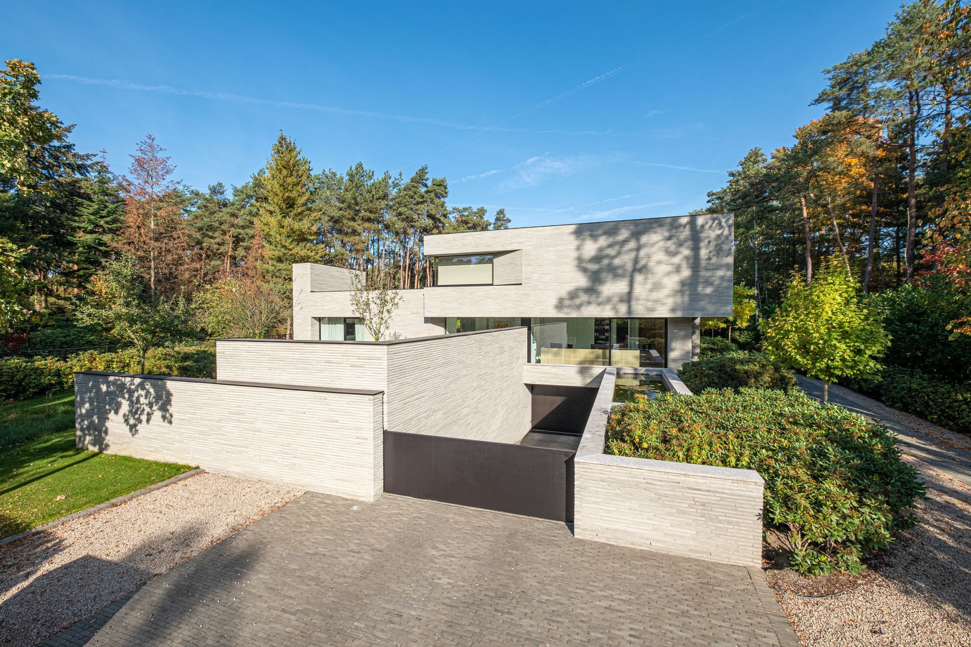 Modern betonnen huis met geometrische terrassen, omgeven door bomen onder een helderblauwe hemel.