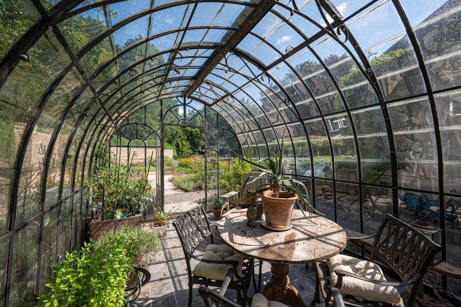 Gebogen metalen kas met planten, een ronde tafel en stoelen in een zonnige tuin.
