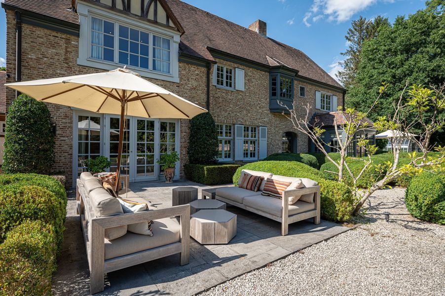 Terrasmeubilair met beige parasol naast een stenen huis en aangelegde tuin.