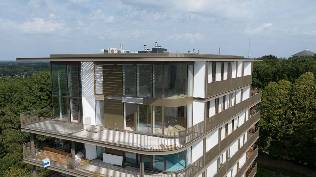 Modern meerlaags gebouw met glazen balkons en een dakterras, omgeven door bomen.
