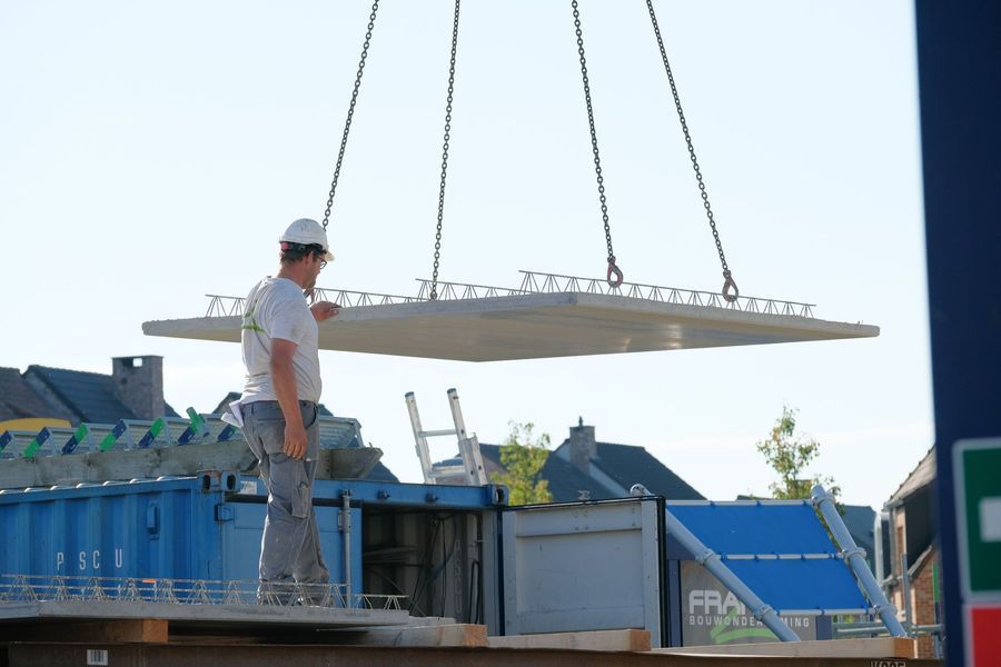 Een arbeider begeleidt een hangend metalen paneel over een blauwe containerwagen op een bouwplaats.