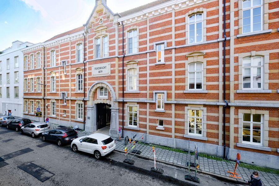 Een gebouw van rode baksteen met witte accenten, een boogvormige ingang en geparkeerde auto's langs een smalle straat.
