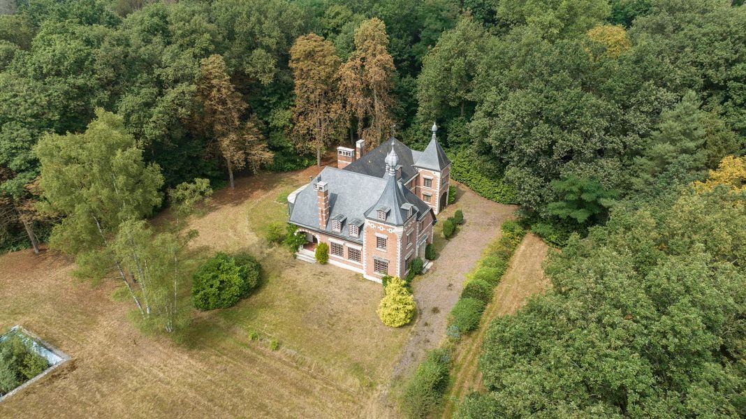 Luchtfoto van een klein grijs huis met een oprit, omgeven door bomen en open grasland.