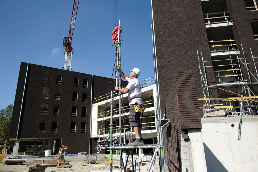 Een arbeider op een steiger installeert apparatuur buiten een bakstenen gebouw in aanbouw.