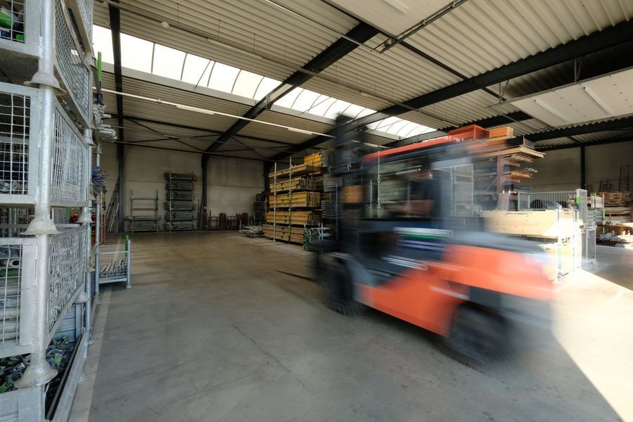 Een oranje heftruck rijdt door een magazijn met gestapelde pallets en stellingen op de achtergrond.