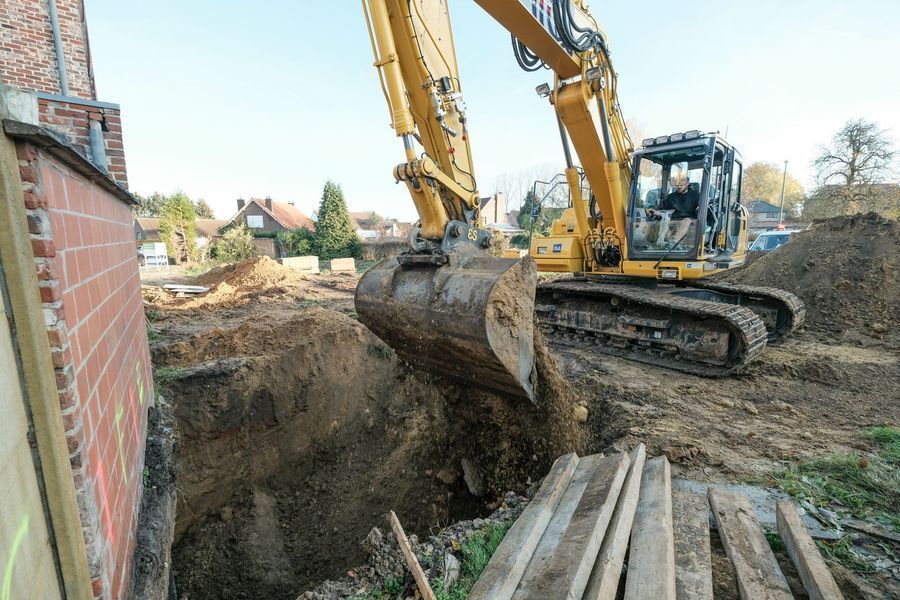 Een graafmachine graaft een sleuf naast een bakstenen gebouw op een modderige bouwplaats.