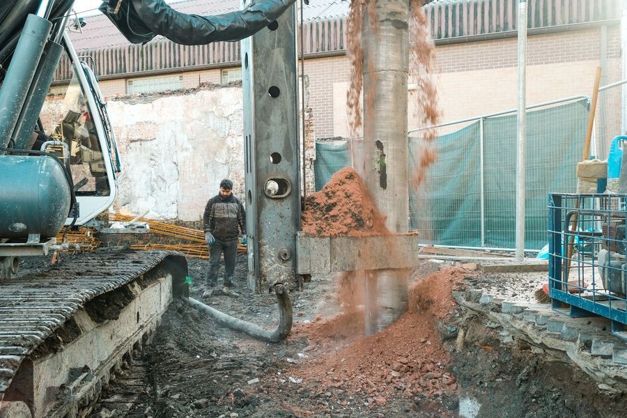 Een graafmachine boort beton naast een arbeider op een bouwterrein, waarbij stof en puin in het rond vliegen.