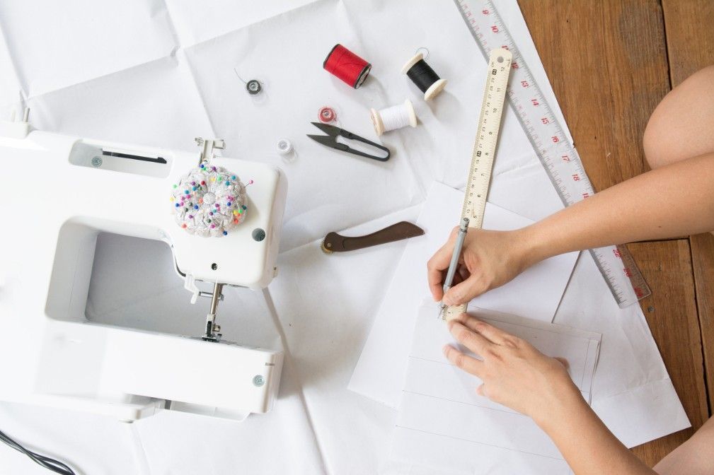 Persona dibujando en papel, máquina de coser, carretes y herramientas sobre una superficie blanca.