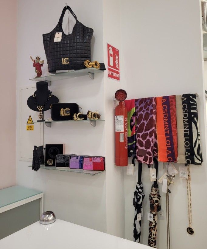 Exhibición de carteras, joyas y bufandas en un ambiente boutique; extintor de incendios y pequeña figura de Jesús visibles.