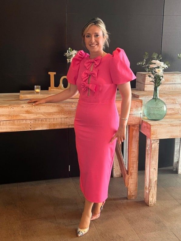 Mujer con vestido rosa con lazos, de pie cerca de una mesa de madera.