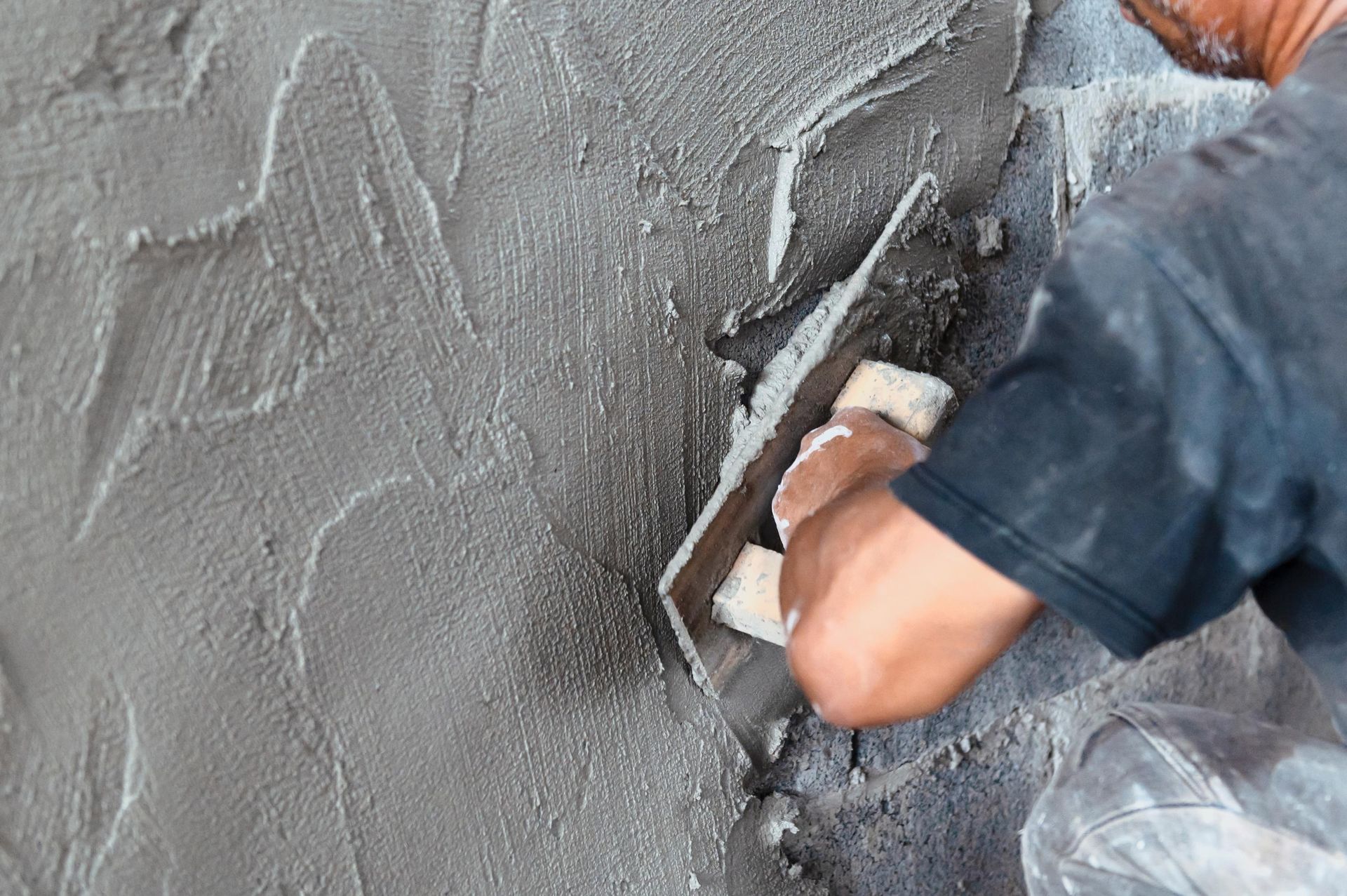 Mann trägt mit einer Kelle Stuck auf eine Wand auf und erzeugt so eine strukturierte graue Oberfläche.