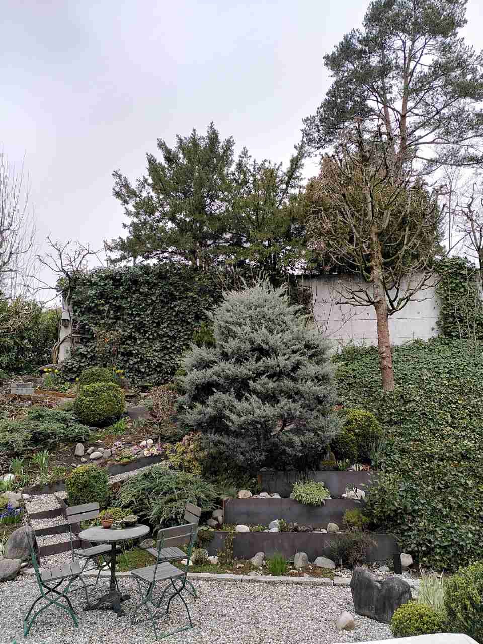 Gartenterrasse mit Steinstufen, Topfpflanzen und einem kleinen Tisch mit Stühlen am Hang.
