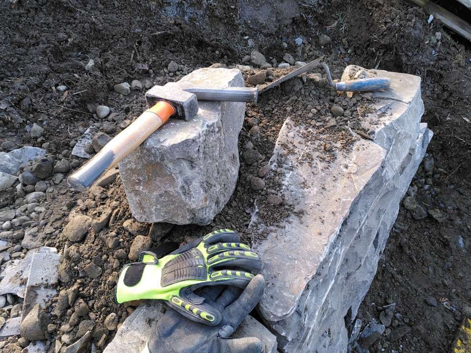 Eine behandschuhte Hand neben einem Felshammer und Meissel auf einem grossen Steinblock im Freien im schlammigen Boden