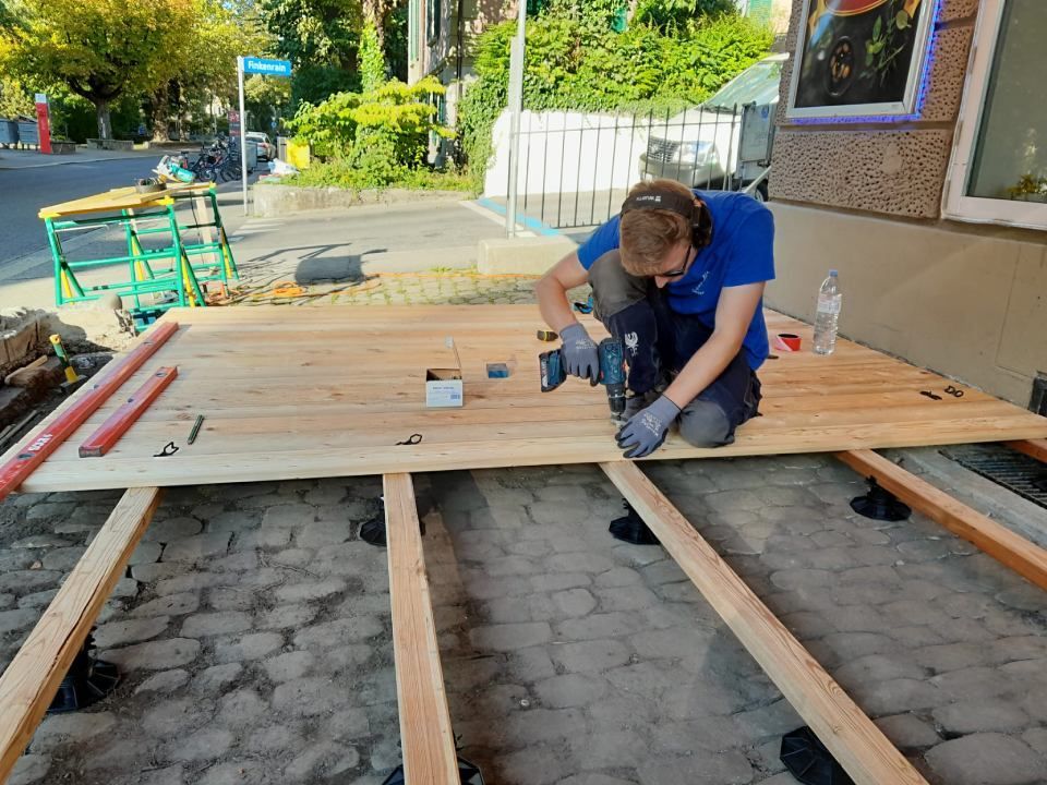 Eine Person kniet im Freien auf einem hölzernen Terrassengerüst und benutzt während Bauarbeiten einen Bohrer.