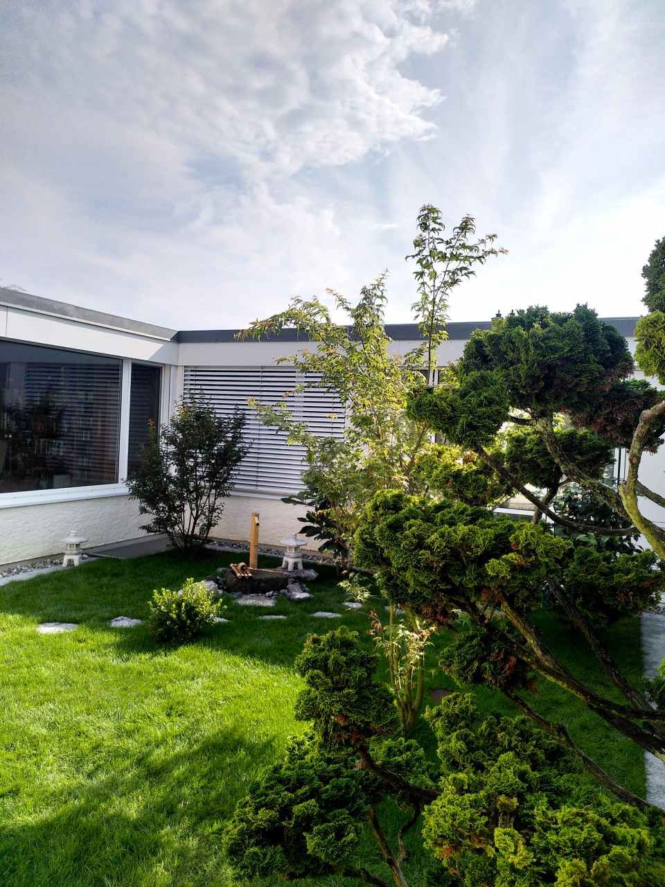 Sonniger Hinterhof mit grünem Rasen, Sträuchern und einem modernen weissen Haus unter einem teilweise bewölkten Himmel