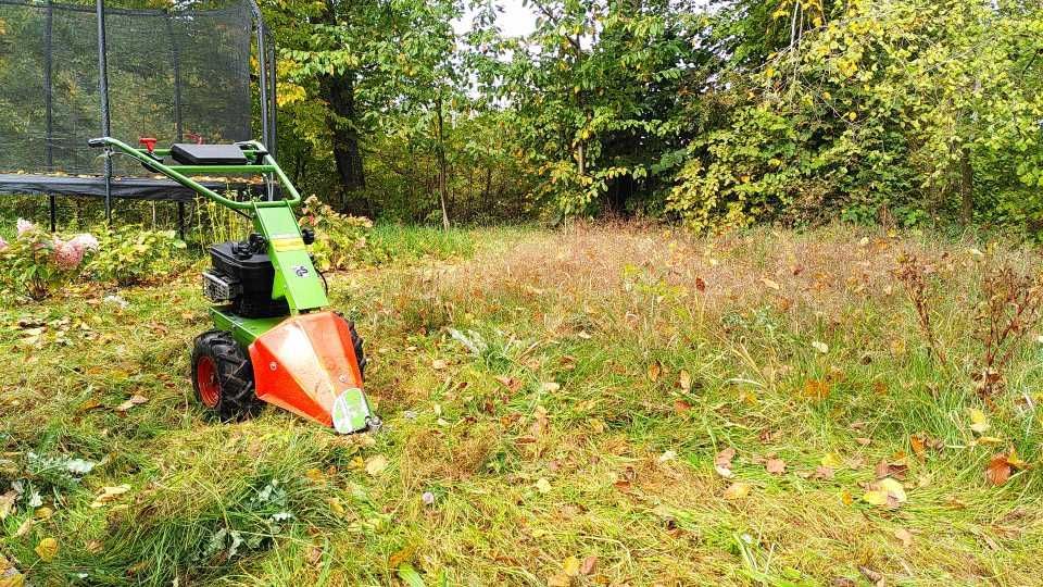 Ein grün-oranger Rasenmäher im hohen Gras nahe Bäumen und Sträuchern.