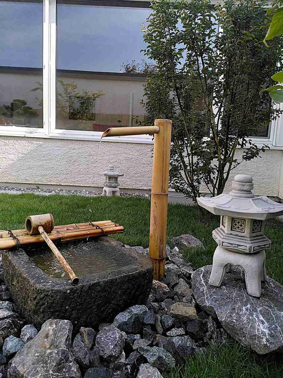 Kleiner japanischer Brunnen im Freien mit Steinen, Bambusrohr und Steinlaterne neben einem Fenster.