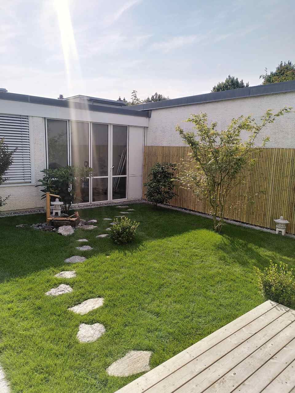 Kleiner, sonniger Hinterhof mit grünem Rasen, Trittsteinen, Terrasse und einer weissen Hausterrasse.