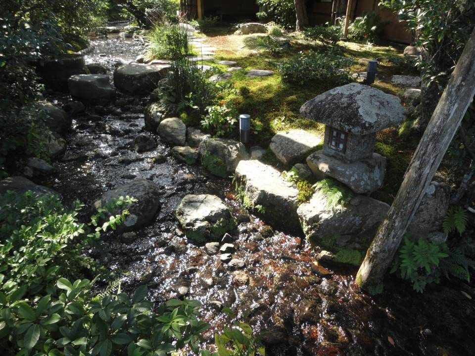 Ein Bachlauf in einem Steingarten mit Trittsteinen, moosbewachsenen Felsen und einer Steinlaterne im gefilterten Sonnenlicht