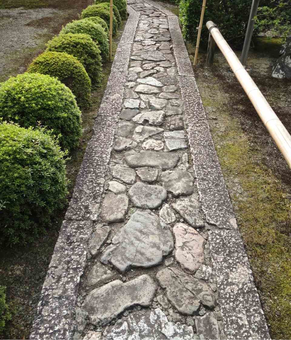 Schmaler Steinweg mit grobem Kopfsteinpflaster, gesäumt von geschnittenen grünen Hecken und einem weissen Geländer.