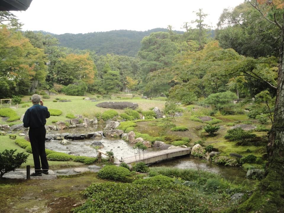 Eine Person steht in einem üppigen japanischen Garten neben einem Steinweg und einem Teich; im Hintergrund erheben sich Hügel.