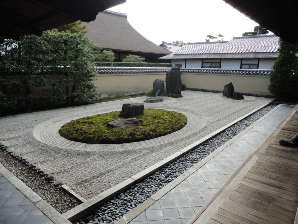 Zen-Garten mit weissem Kies, Moosinsel, Steinlaternen und Holzsteg neben einem traditionellen Gebäude.
