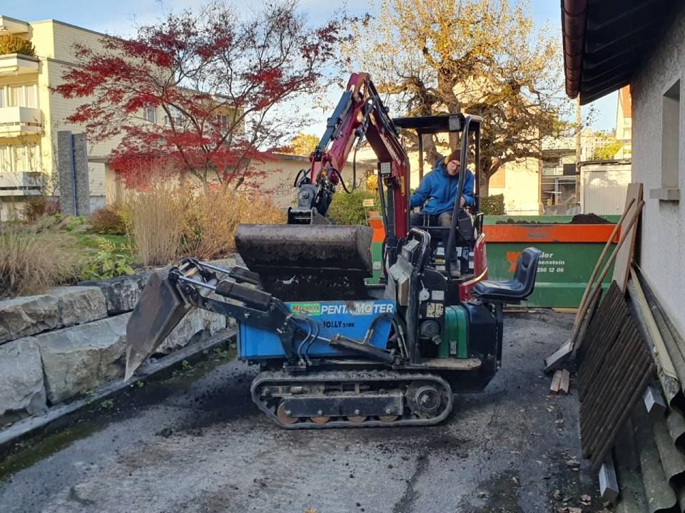 Ein Minibagger und ein Arbeiter reparieren eine Einfahrt neben einem Haus im Herbstlaub.