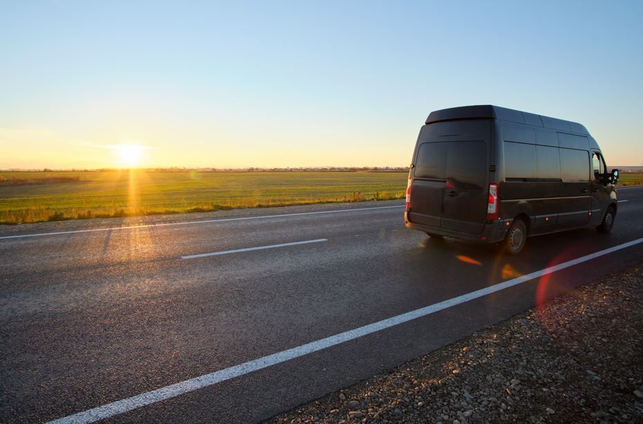 Furgoneta negra circulando por una carretera asfaltada al atardecer, con campos al fondo.