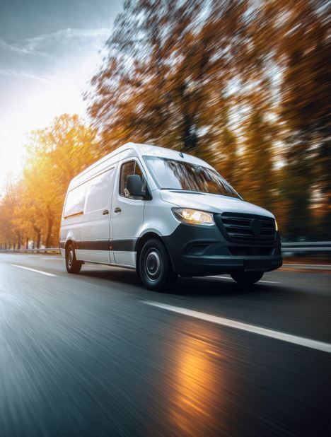Furgoneta blanca circulando a toda velocidad por una carretera con un fondo borroso de árboles otoñales y sol poniente.