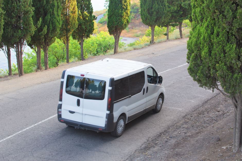 Furgoneta blanca circulando por una carretera bordeada de árboles, cerca del agua.