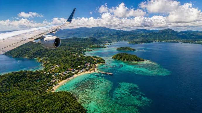 Ala de avión sobre una isla tropical con aguas turquesas, playas de arena y colinas verdes.