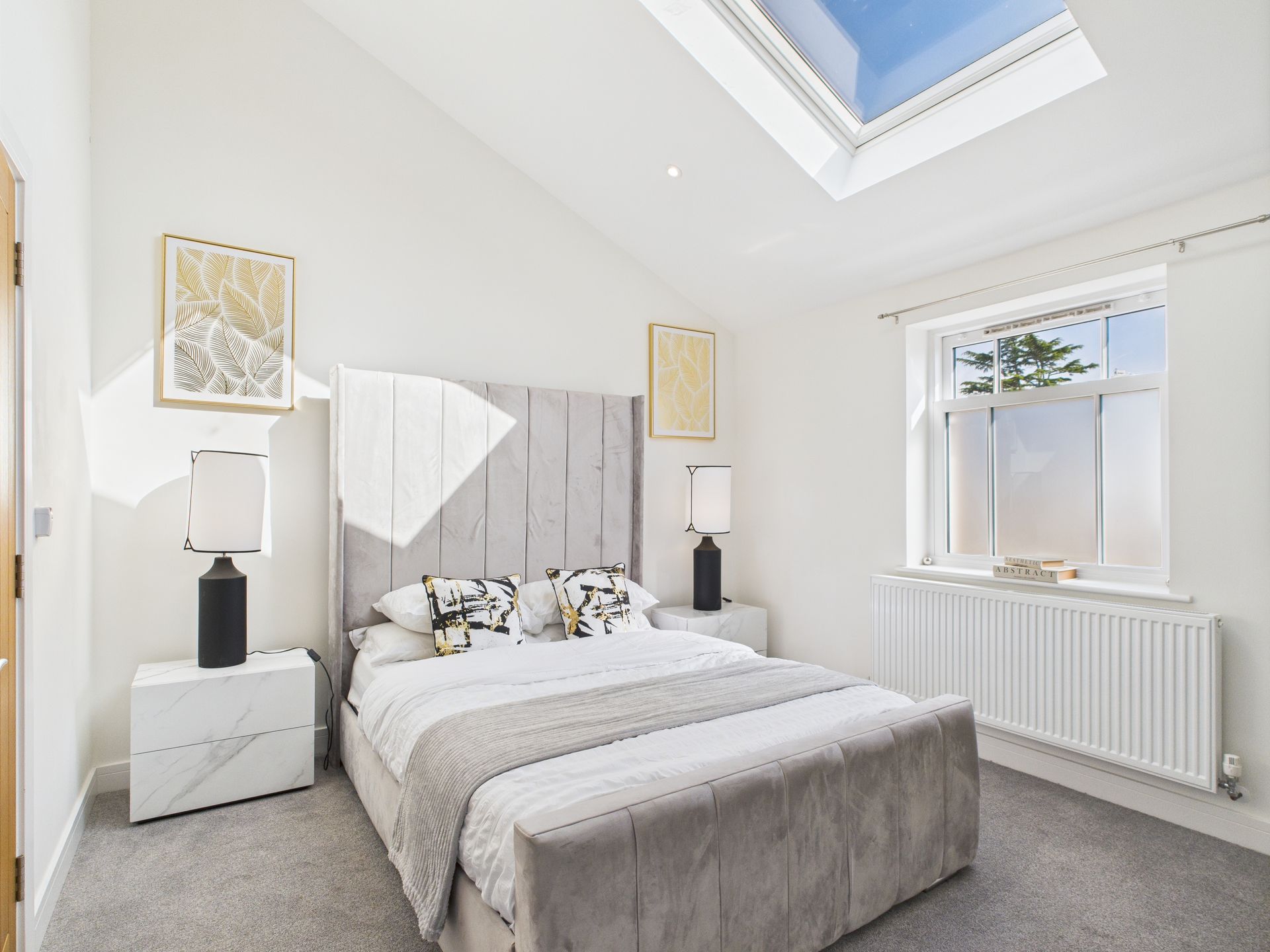 A bright bedroom with a grey tufted bed, white nightstands, patterned pillows, a skylight, and a radiator under a window.