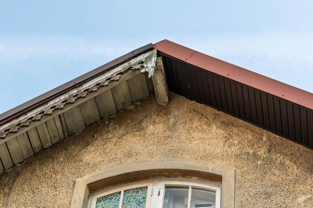 Coin d'un bâtiment au toit brun et à la façade en stuc beige patiné ; un ciel bleu est visible.