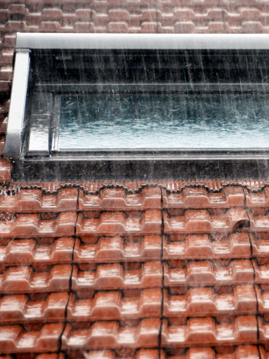 Pluie tombant sur un toit de tuiles rouges percé d'une lucarne rectangulaire.